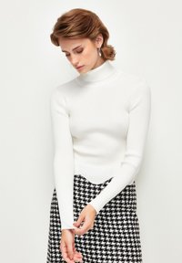 Biała sweterka z golfem, z długimi rękawami, z falbaniastym dołem, w połączeniu z czarną i białą spódnicą w wzór houndstooth.