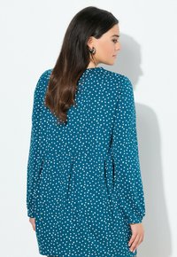 Vrouw met lang donker haar, gekleed in een teal jurk met witte driehoekpatronen, die zijwaarts staat tegen een effen witte muur.