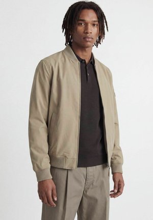 Ung mand med skulderlange dreadlocks iført en beige bomberjakke, sort strikket polo og matchende beige bukser mod en ensfarvet baggrund.