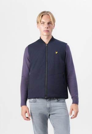 Veste matelassée marine avec fermeture éclair à l'avant, dotée d'un petit logo jaune sur la poitrine gauche, portée sur une chemise à manches longues violette.