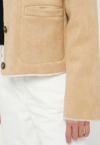 Beżowa zamszowa kurtka o fakturowanym wykończeniu, z kieszenią na piersi, okrągłymi guzikami i puszystymi mankietami, zestawiona z białymi spodniami.