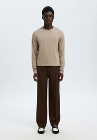 Νεαρός άνδρας που στέκεται, φορώντας μπεζ πουλόβερ με μακριά μανίκια, σκουρόχρωμο καφέ παντελόνι φαρδιάς γραμμής και μαύρα-λευκά loafers σε απλό ανοιχτό φόντο.