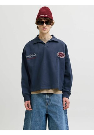 Noor inimene, kes kannab burgundia värvi mütsi, tumedaid päikeseprille, tumesinist triiksärki plaastritega, beeži aluspluusi ja laia helesinist pükse, seistes.