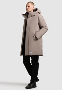 Bézs, térdig érő steppelt kabát kapucnival, két oldalzsebével és logó foltjával az alsó részen, fekete nadrággal és cipővel párosítva.