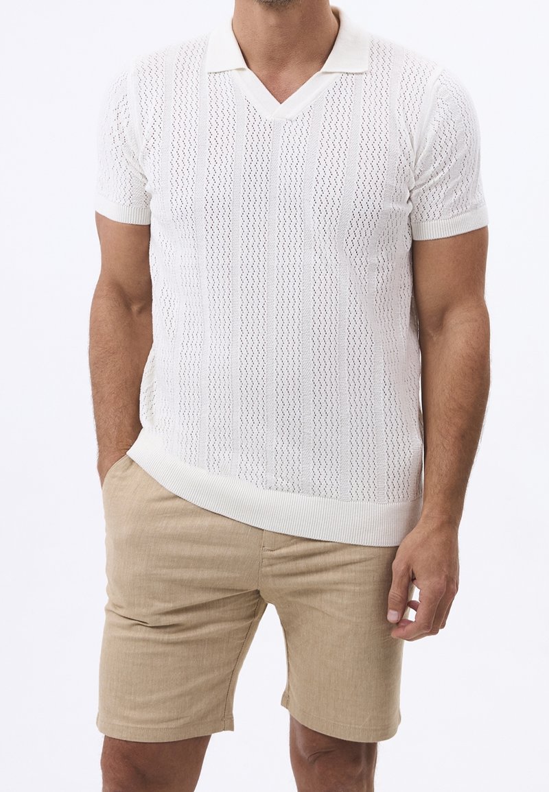 Mand iført hvid kortærmet strik polo skjorte med tekstureret mønster og beige knælange shorts, hænderne afslappet ved siden.