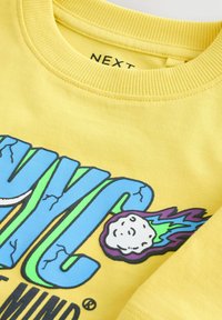 Gul bomulds sweatshirt med et blåt og grønt grafikdesign, der har en karakter og flammeakcenter, ribbet rund hals og manchetter.