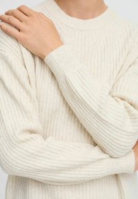 Pull en maille côtelée beige avec un col rond et des manches longues. Présente un tissu texturé et des poignets ajustés. Design simple, sans motifs supplémentaires.