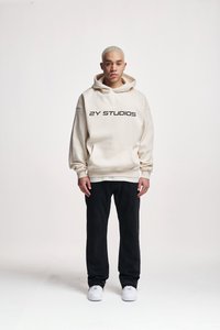 Hanorac crem cu "2Y STUDIOS" imprimat în negru, asortat cu pantaloni negri și sneakers albi. Detalii vizibile ale glugii și buzunarului din față.