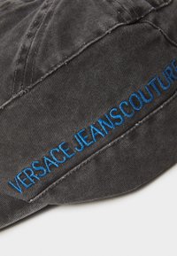 Casquette en tissu gris avec le texte brodé bleu "VERSACE JEANSCOUTURE." Texture usée avec des coutures visibles et un design décontracté.