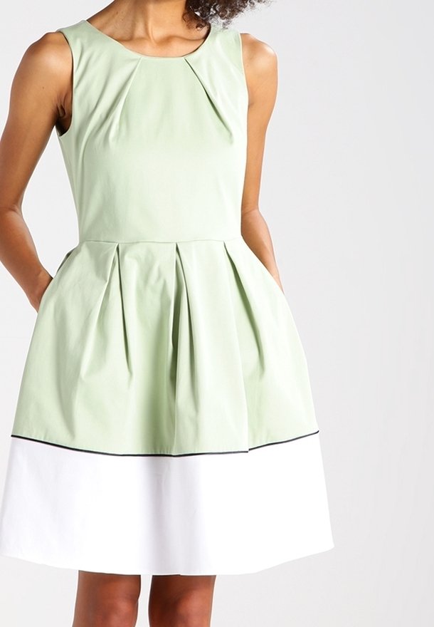 Robe verte sans manches avec des plis à la taille et une jupe blanche. Elle présente un col rond et un ourlet bordé de noir. Texture lisse.
