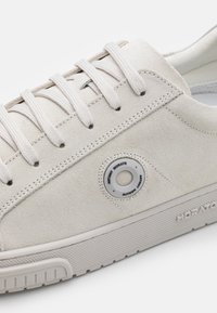 Baskets en suède blanc avec un détail de logo rond, des lacets plats et une semelle en caoutchouc texturée. Design simple avec des accents minimalistes.