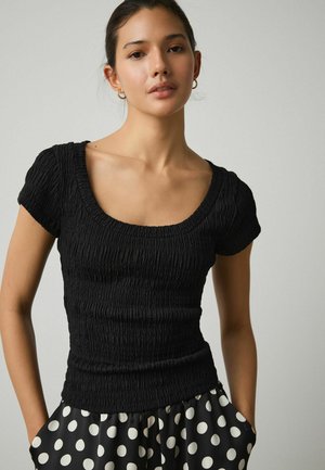 Ung kvinde med mørkt hår iført en sort, tekstureret top med korte ærmer og sorte bukser med store hvide polkaprikker, stående med hænderne i lommerne.