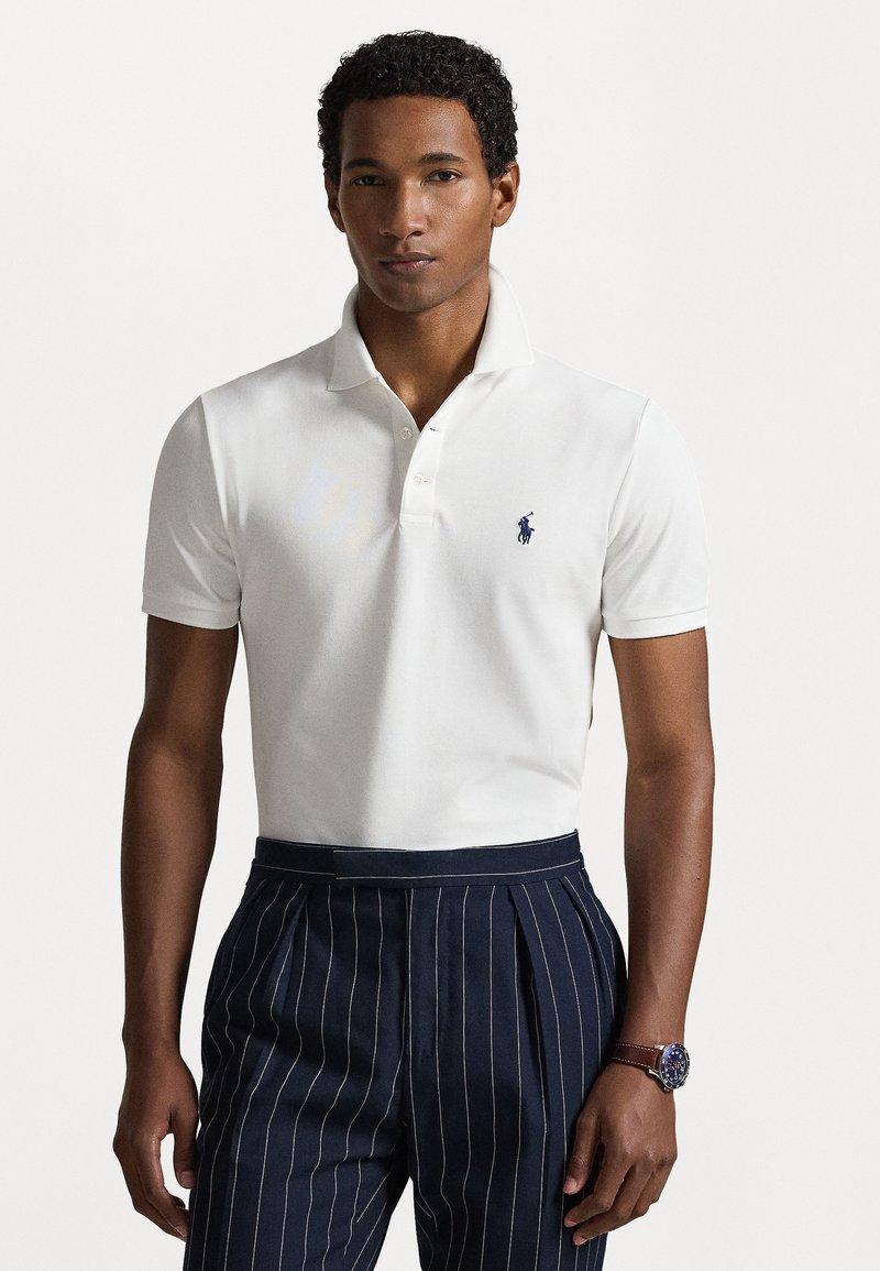 Hvit polo skjorte i bomull med en to-knapp placket og marineblå brodert logo. Kombinert med marineblå stripete bukser og et klokke.