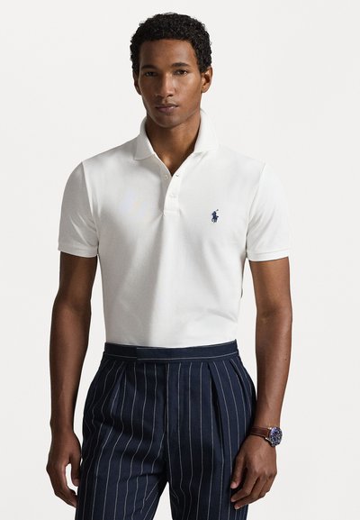Biela polo košeľa z bavlny s dvojitou gombíkovou léga a vyšitým logom v námorníckej modrej farbe. V kombinácii s námorníckymi pruhovanými nohavicami a hodinkami.