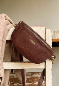 Brązowa torba ze skóry z tłoczeniem w motyw krokodyla, z suwakiem i złotymi akcentami, z odpinanym paskiem oraz kompaktowym, zaokrąglonym kształtem.