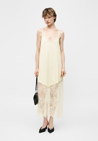 Femeie purtând o rochie crem tip slip cu dantelă suprapusă, ținând o mică geantă neagră și purtând pantofi negri ascuțiți, stând în fața unui fundal simplu.