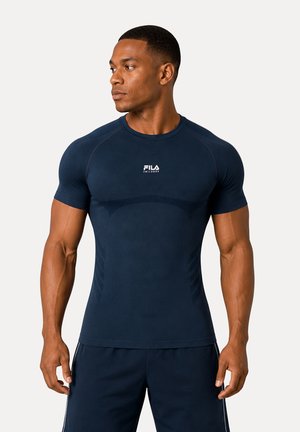 Athletischer Mann, der nach links schaut, trägt ein eng anliegendes dunkelblaues FILA-Kurzarmshirt und passende Shorts vor einfachem Hintergrund.