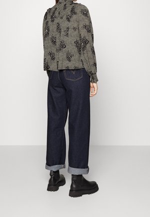 Woman wearing a high-neck floral blouse, dark cuffed wide-leg jeans, and black chunky ankle boots, standing against a plain background.