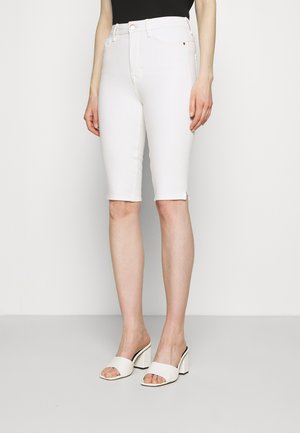 Kvinde iført hvide knælange tætsiddende shorts, sort top og hvide åbne blokhæle-sandaler, stående mod en ensfarvet baggrund.