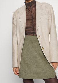 Beige blazer met twee knopen, gecombineerd met een doorzichtige bruine top met hoge hals en een textuur rok in olijfgroen. Zachte stof met subtiele details.