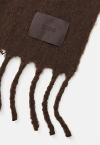 Brown wool scarf featuring fringed edges and a rectangular dark label with embossed text. Soft texture with a fuzzy appearance.