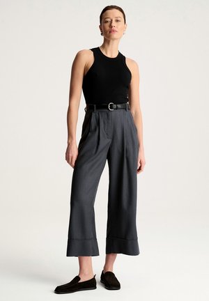 Kobieta stoi, ubrana w czarny top bez rękawów, szare szerokie spodnie z wysokim stanem i paskiem oraz czarne mokasyny na jednolitym tle.