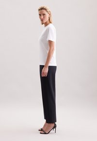 Wit T-shirt met korte mouwen gecombineerd met zwarte, op maat gemaakte broek en zwarte sandalen met hoge hak, gepresenteerd tegen een neutrale achtergrond.