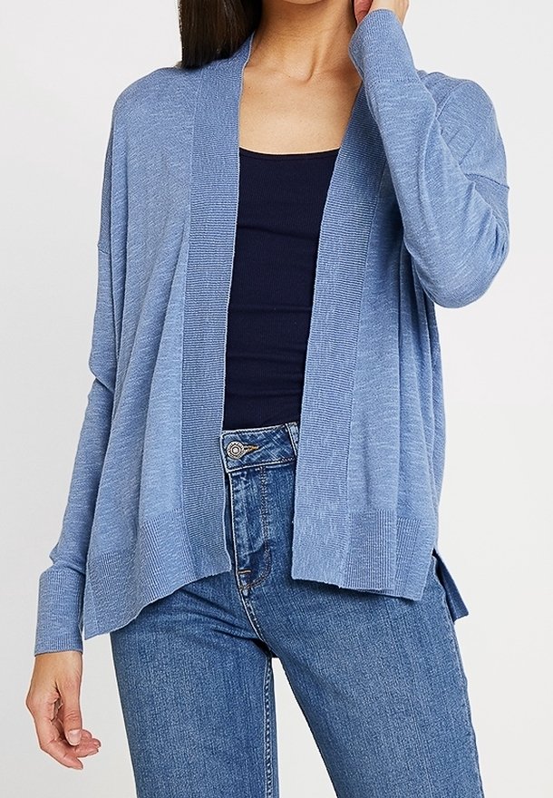 Femme portant un cardigan bleu ample sur un haut noir et un jean bleu, avec une main touchant ses cheveux près du visage.