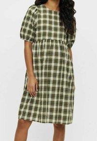 Frau trägt ein knielanges, grün-weiß kariertes Kleid mit gepufften Kurzärmel und einer gerafften Taille.