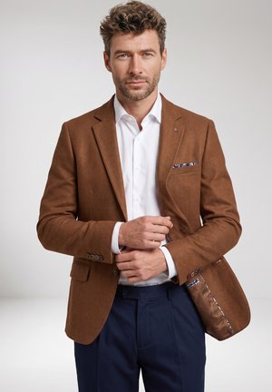Man with curly hair wearing a brown blazer, white shirt, and navy pants, adjusting his blazer with a neutral background.