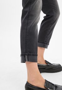 Jambes inférieures portant un jean noir retroussé avec des boutons décoratifs sur les côtés et des mocassins noirs brillants sur fond blanc.