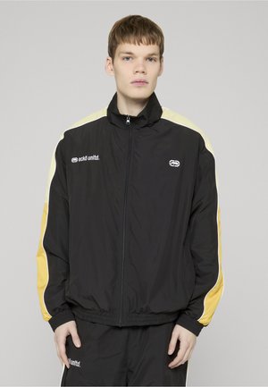 Mladý muž oblečený v čiernej bunde na zips a tmavých nohaviciach so žltými pruhmi, stojaci pred jednoliatym svetlým pozadím.