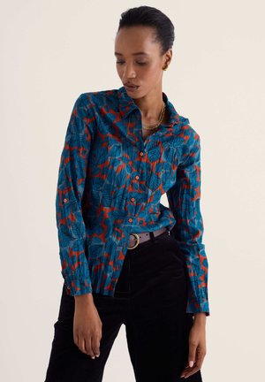 Femme portant une chemise boutonnée à motif de feuilles bleu et rouge avec un pantalon noir, regardant vers le bas sur un fond beige uni.