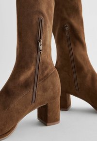 Bottes hautes jusqu'au genou en daim marron avec un talon bloc, dotées de fermetures éclair latérales et de détails surpiqués pour plus de texture et de design.