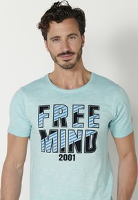Világoskék, rövid ujjú póló, amelyen vastag fekete és kék csíkos betűkkel olvasható a "FREE MIND" felirat. A póló kerek nyakkivágással és puha tapintással rendelkezik.