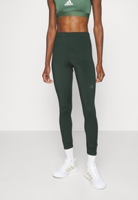 Zelené legíny s elastickým designem, hladkou texturou a manžetami na kotnících, doplněné o odpovídající zelenou sportovní podprsenku a bílé sportovní boty.