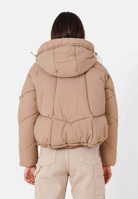 Giacca a vento in beige chiaro, con cappuccio regolabile, texture trapuntata e polsini elasticizzati. Abbinata a pantaloni cargo di colore chiaro.