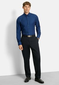 Mann, der ein tailliertes blaues Hemd, schwarze Hosen mit Gurt und schwarze Lederschuhe trägt, vor einem weißen Hintergrund steht.