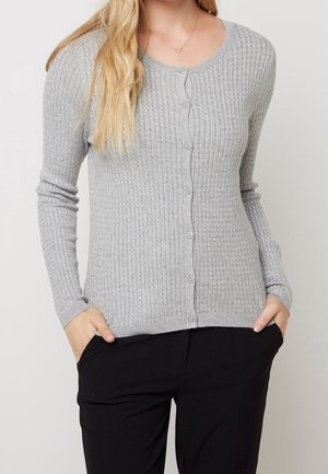 Femme portant un cardigan boutonné côtelé gris clair à manches longues et un pantalon noir, les mains dans les poches, debout devant un fond uni.