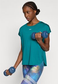 Κοντομάνικο αθλητικό τοπ σε τιρκουάζ με στρογγυλή λαιμόκοψη, φορεμένο πάνω από πολύχρωμο κολάν με λουλουδένια σχέδια και διακοσμητικά από δίχτυ. Κρατά μπλε αλτήρες.