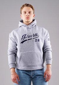 Junger Mann steht, trägt einen hellgrauen Kapuzenpullover mit dem Text "Picardi 28" und eine blaue Jeans vor einem schlichten grauen Hintergrund.