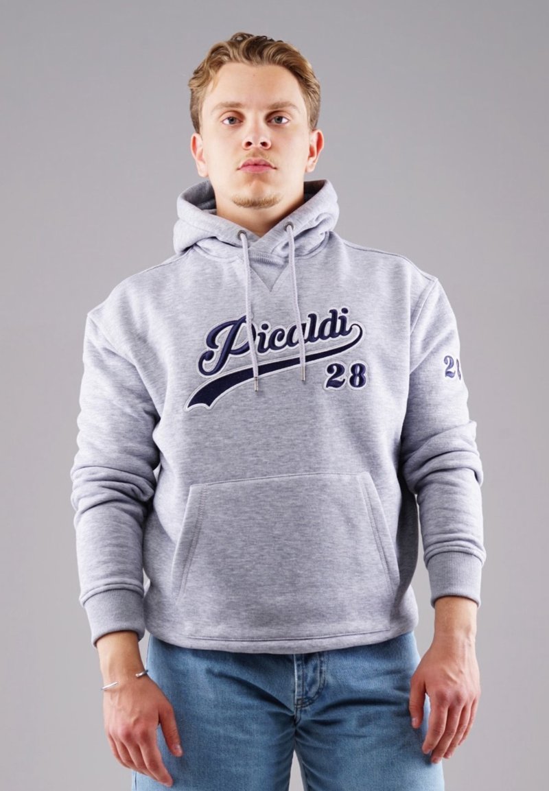Junger Mann steht, trägt einen hellgrauen Kapuzenpullover mit dem Text "Picardi 28" und eine blaue Jeans vor einem schlichten grauen Hintergrund.