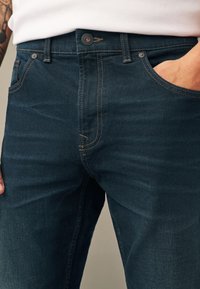 Dunkelblaue Denim-Jeans mit sichtbaren Nähten, vorderem Knopf und einer teilweise in der rechten Tasche steckenden Hand vor einem weißen Hemd.