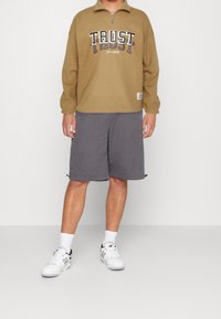 Brun sweatshirt med lynlås og teksten "TRUST", kombineret med grå shorts. Modellen bærer hvide sneakers og sokker og udstråler en afslappet stil.
