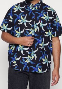 Chemise à manches courtes avec boutons, noire avec un motif floral comprenant des fleurs bleues, blanches et jaunes. Matière en coton, coupe décontractée.
