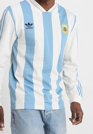 Mężczyzna noszący długi rękaw koszulkę piłkarską Argentyny w niebiesko-białe pionowe paski, z logo Adidas i herbem AFA.