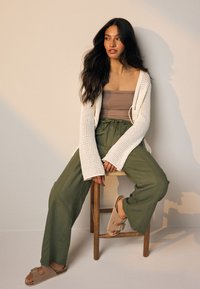 Cardigan tricotat crem, top bej, pantaloni largi kaki și șlapi de culoare bronz. Model așezat pe un scaun de lemn, pe un fundal deschis.