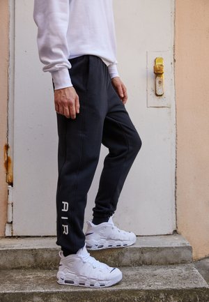 Person, der schwarze Sweatpants mit weißem "AIR"-Schriftzug und weiße Nike-Sneaker trägt, steht auf Betontreppen in der Nähe einer weißen Tür mit rostigem Schloss.