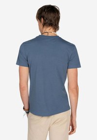 Camiseta de manga corta en suave algodón azul. Presenta un cuello redondo y un diseño sencillo. Llevada con pantalones cortos de un beige claro, vista desde atrás.