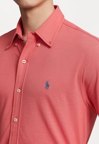 Korálová polo košile z texturované bavlny, s klasickým límcem, zapínáním na knoflíky a modrou vyšitou nášivkou na levé hrudi.